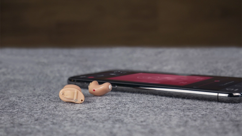 Dos audífonos intracanales sobre una mesa junto a un teléfono móvil: cada uno es más pequeño que la yema de un dedo